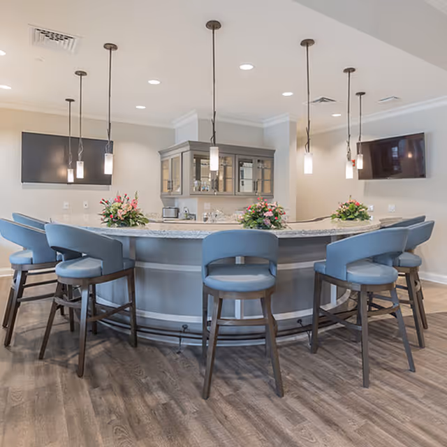 Curved communal bar area with blue upholstered stools, pendant lights, floral arrangements, and two wall-mounted TVs.