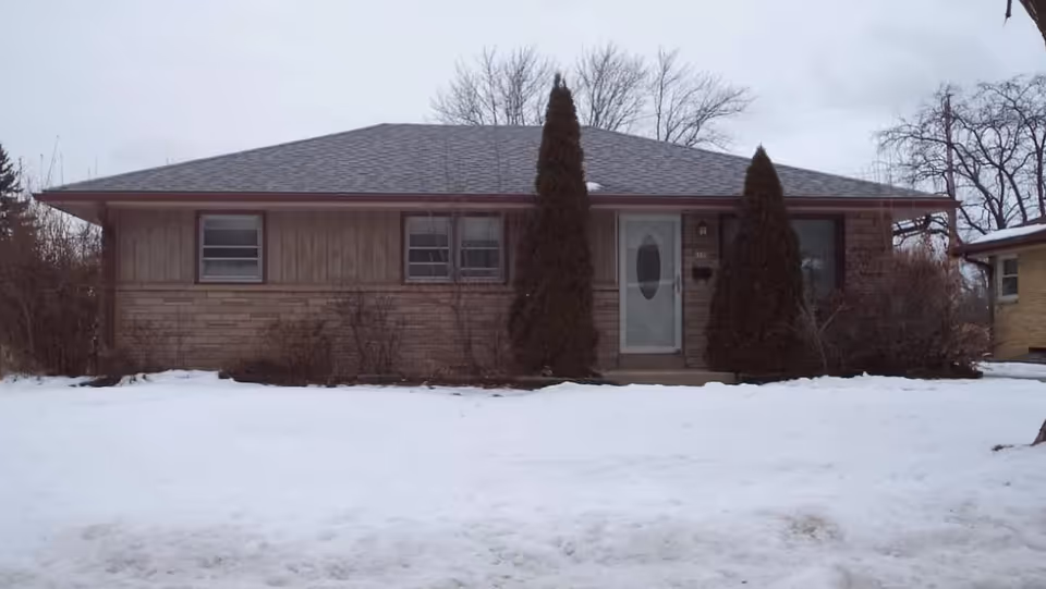 Single-story brick and wood house with a central front door flanked by two tall evergreen shrubs and a snow-covered lawn.
