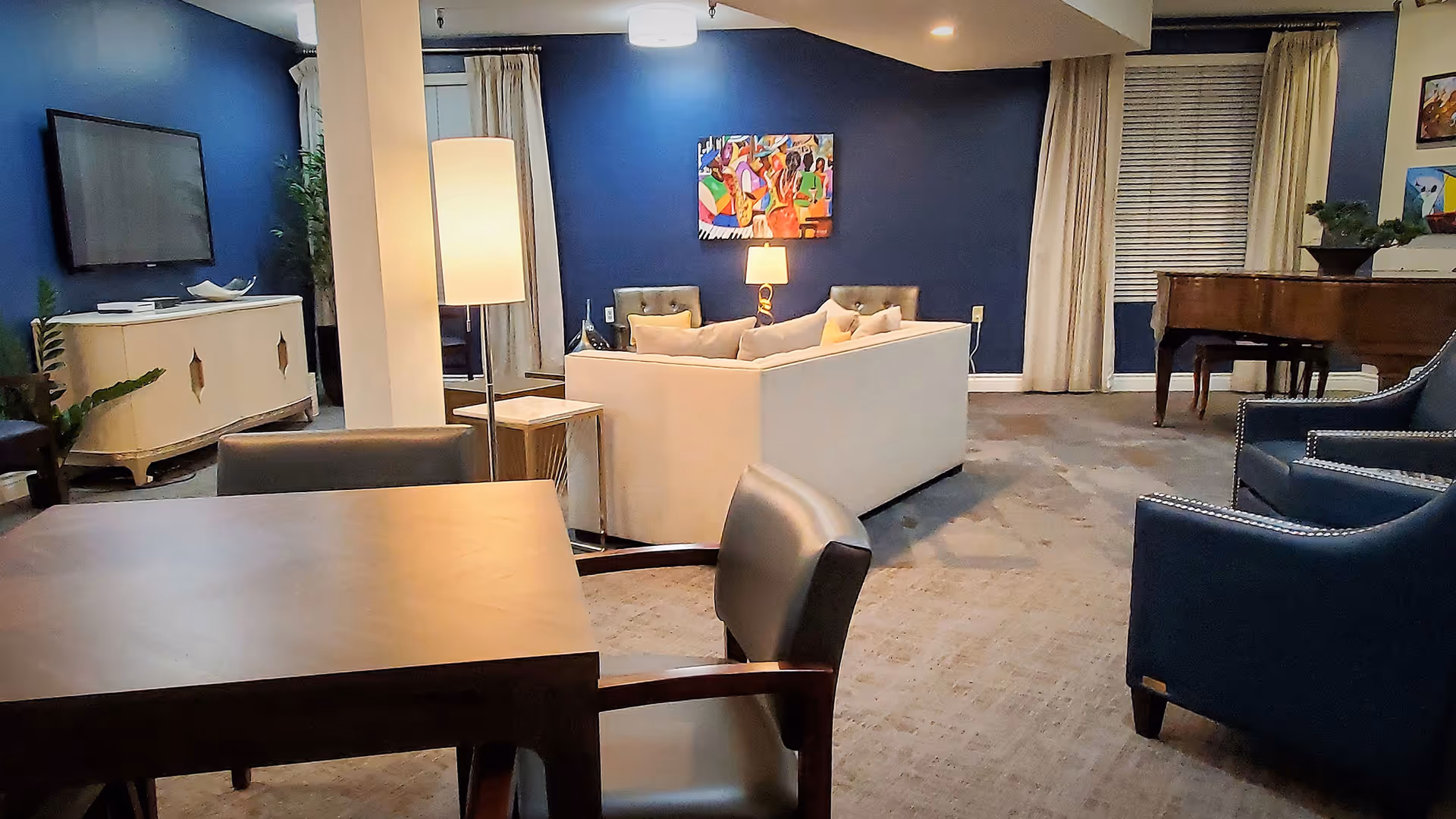 Community lounge with sofas, armchairs, a TV, piano and tables in a senior living facility.