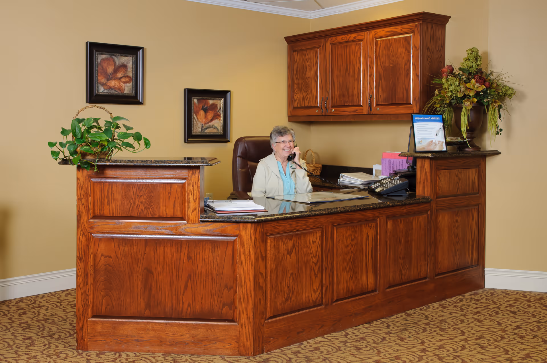 Reception area with a wooden desk and a smiling woman sitting behind it, talking on the phone. The desk has a plant, office supplies, and a sign. The wall behind is beige with two framed floral pictures and wooden cabinets above the desk.