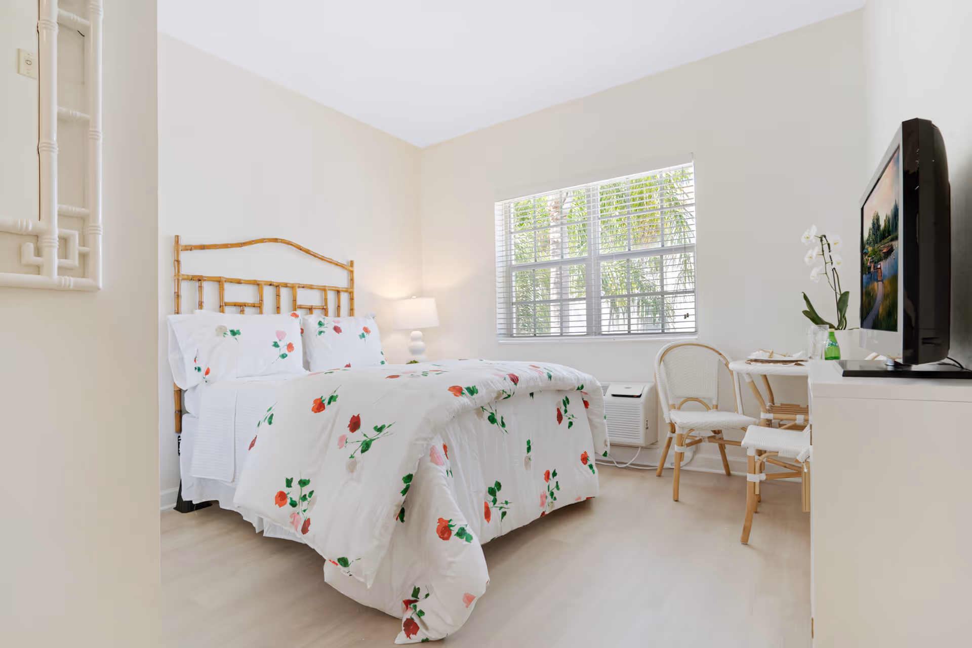 Sunlit bedroom with a floral comforter, bamboo headboard, window blinds, TV on a dresser and a small table with chairs.