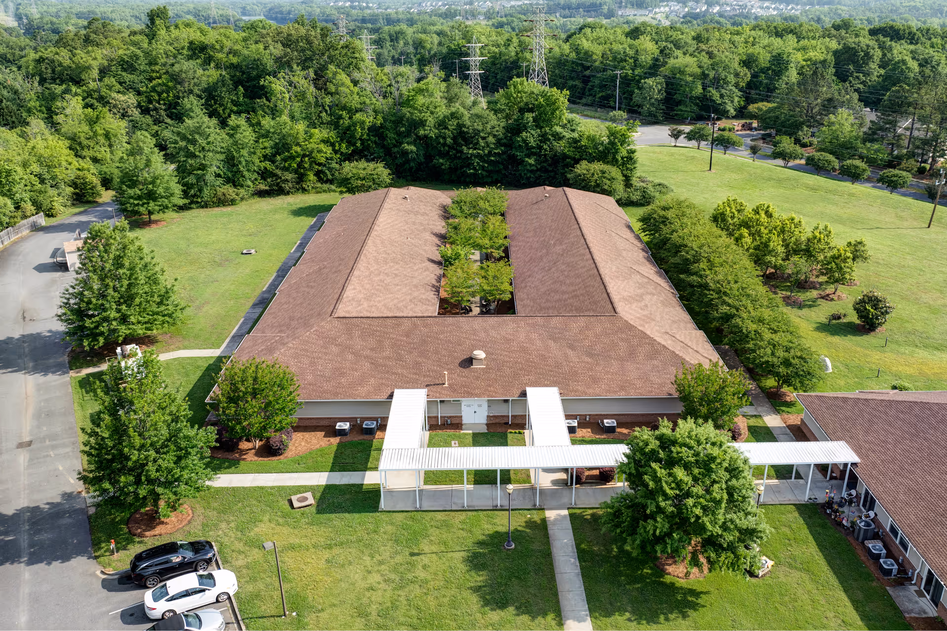 Aerial view of a single-story building with a brown roof surrounded by green lawns and trees. The building has a covered walkway leading to the entrance and is situated in a spacious, wooded area with a parking lot visible on the left side.