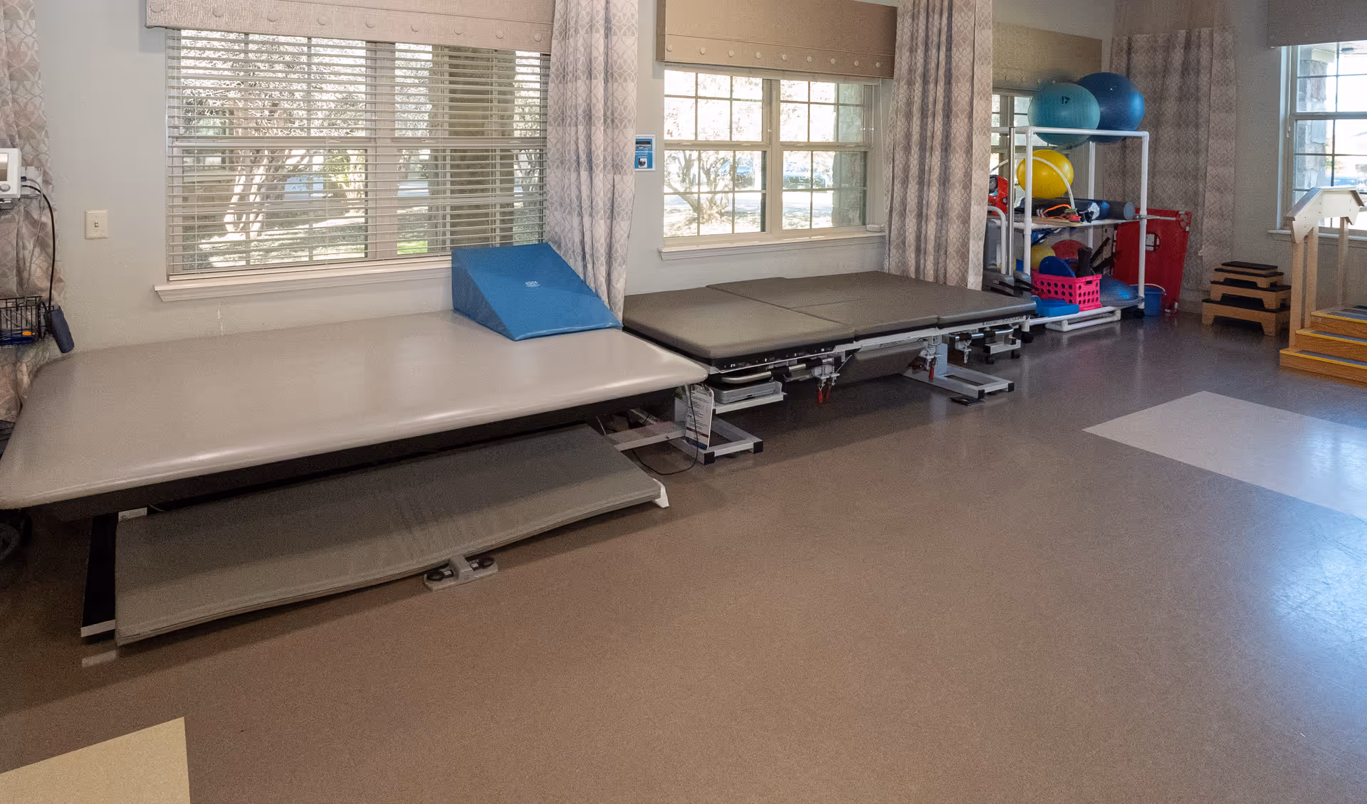 A therapy or rehabilitation room with two padded treatment tables positioned against windows with blinds and curtains. There are exercise balls and other physical therapy equipment on shelves in the corner, and a set of stairs with handrails is visible on the right side.
