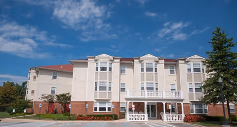 Exterior view of a three-story senior living facility building with beige siding and red brick accents under a blue sky with scattered clouds. The building has multiple windows, a covered entrance with white railings, and landscaped bushes and trees around the parking area.