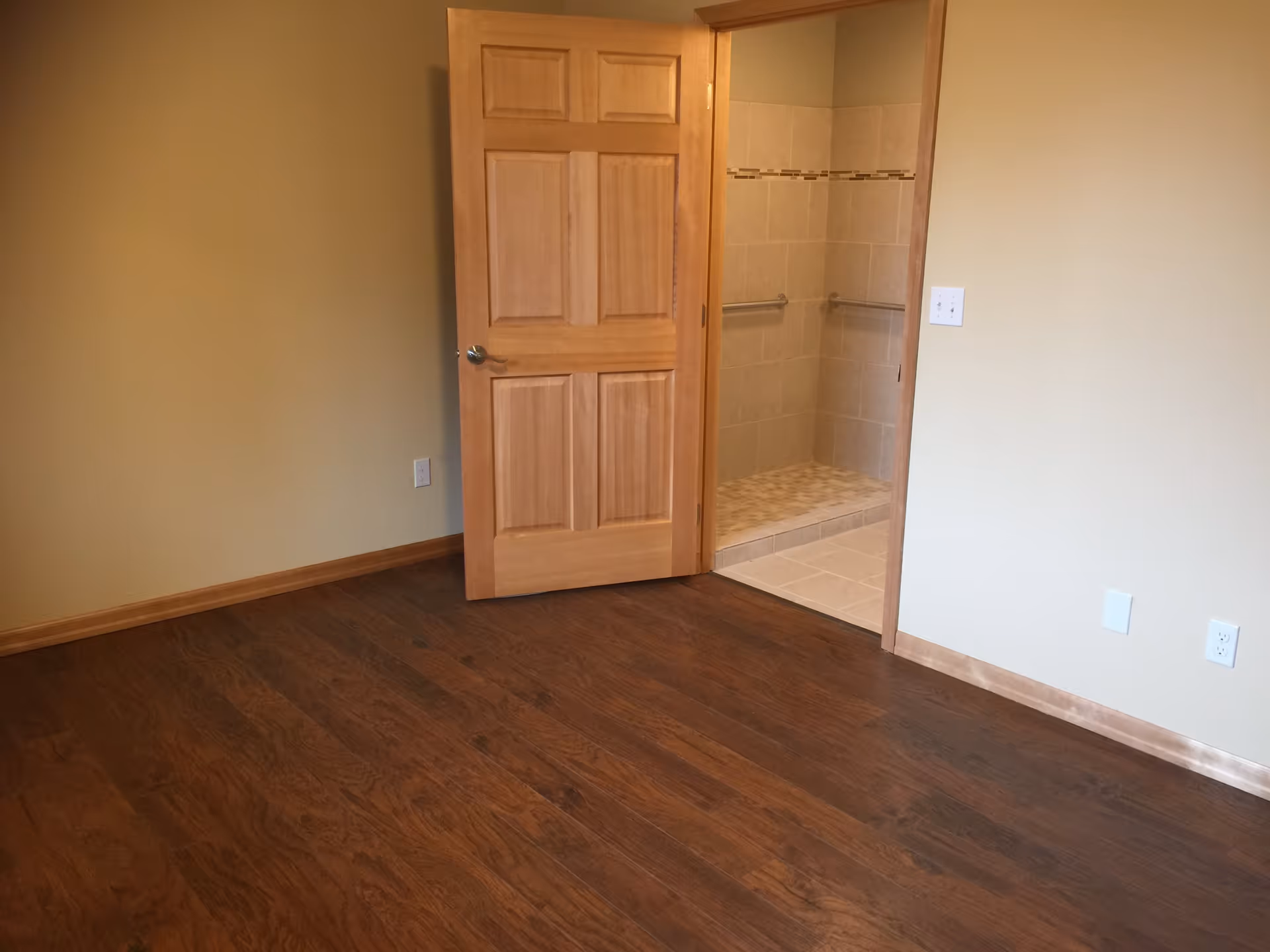 Empty room with wooden floor and beige walls, featuring an open wooden door leading to a tiled bathroom with grab bars installed on the walls.