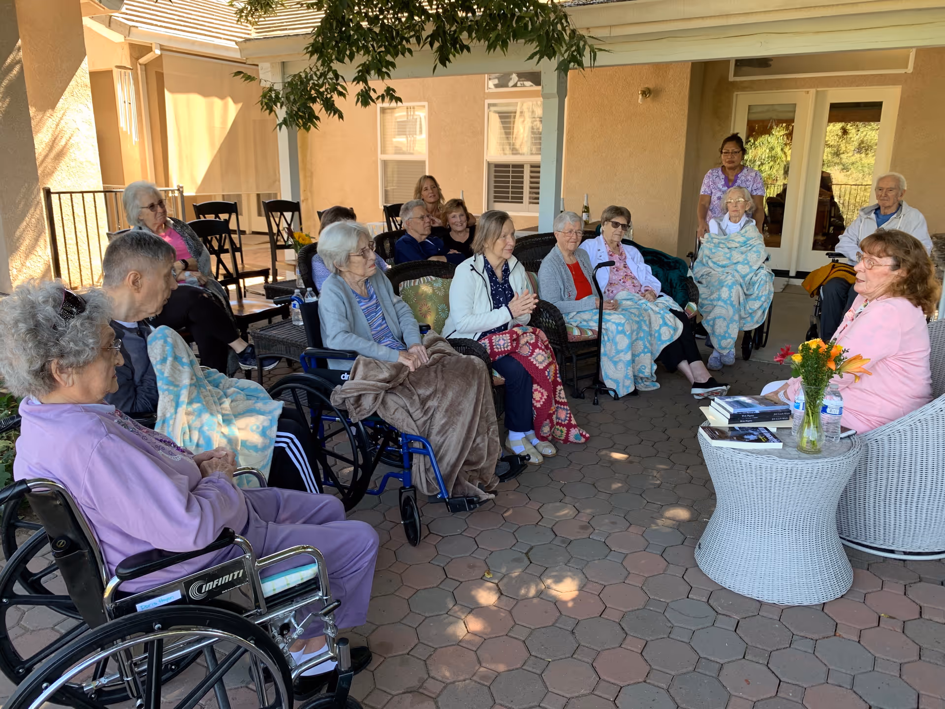 A group of elderly people sitting in a semi-circle on a covered patio, some in wheelchairs and others in chairs, many with blankets. A woman in a pink jacket is seated at a white wicker table with flowers and books, engaging with the group. The setting is outdoors with a building visible in the background.