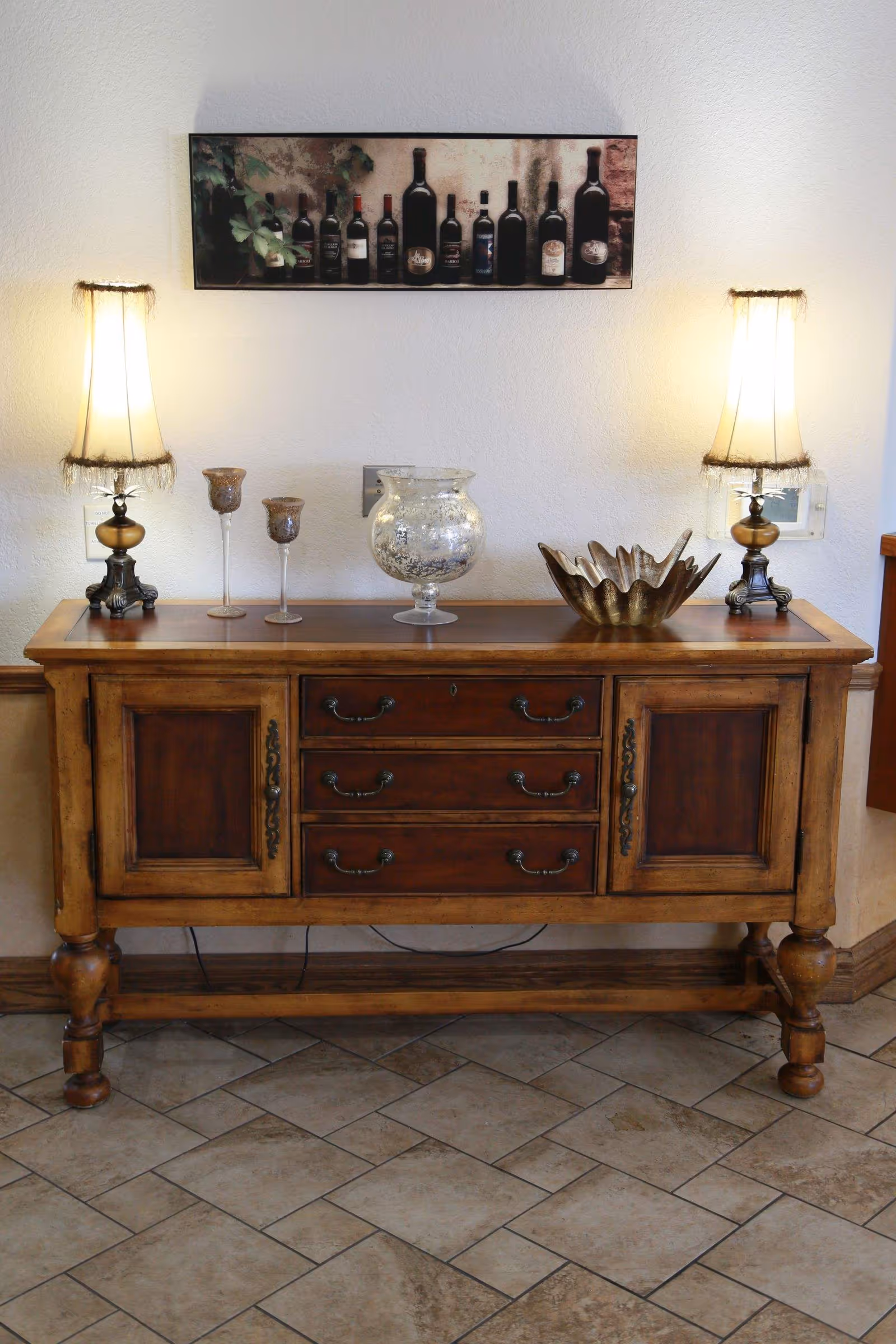 A wooden sideboard with three drawers and two cabinets, decorated with two vintage-style lamps with fringed shades, two candle holders, a glass vase, and a decorative bowl. Above the sideboard is a wall art piece featuring various wine bottles. The floor is tiled with a beige and brown pattern.