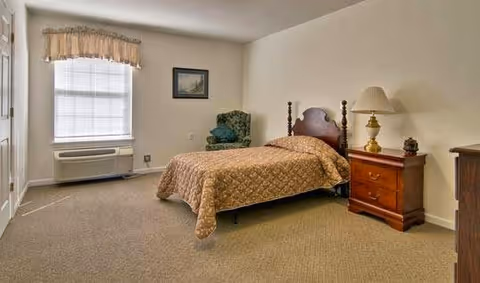 A simple furnished bedroom with a single bed, wooden nightstand and lamp, upholstered chair, window with valance, and beige carpet.