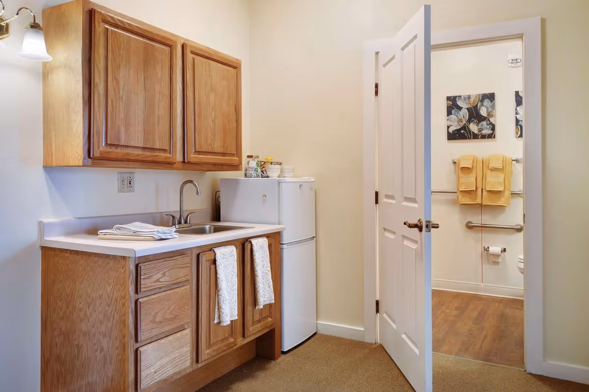 Small kitchenette with wooden cabinets, a sink and mini refrigerator, and an open door revealing a bathroom with grab bars and yellow towels.