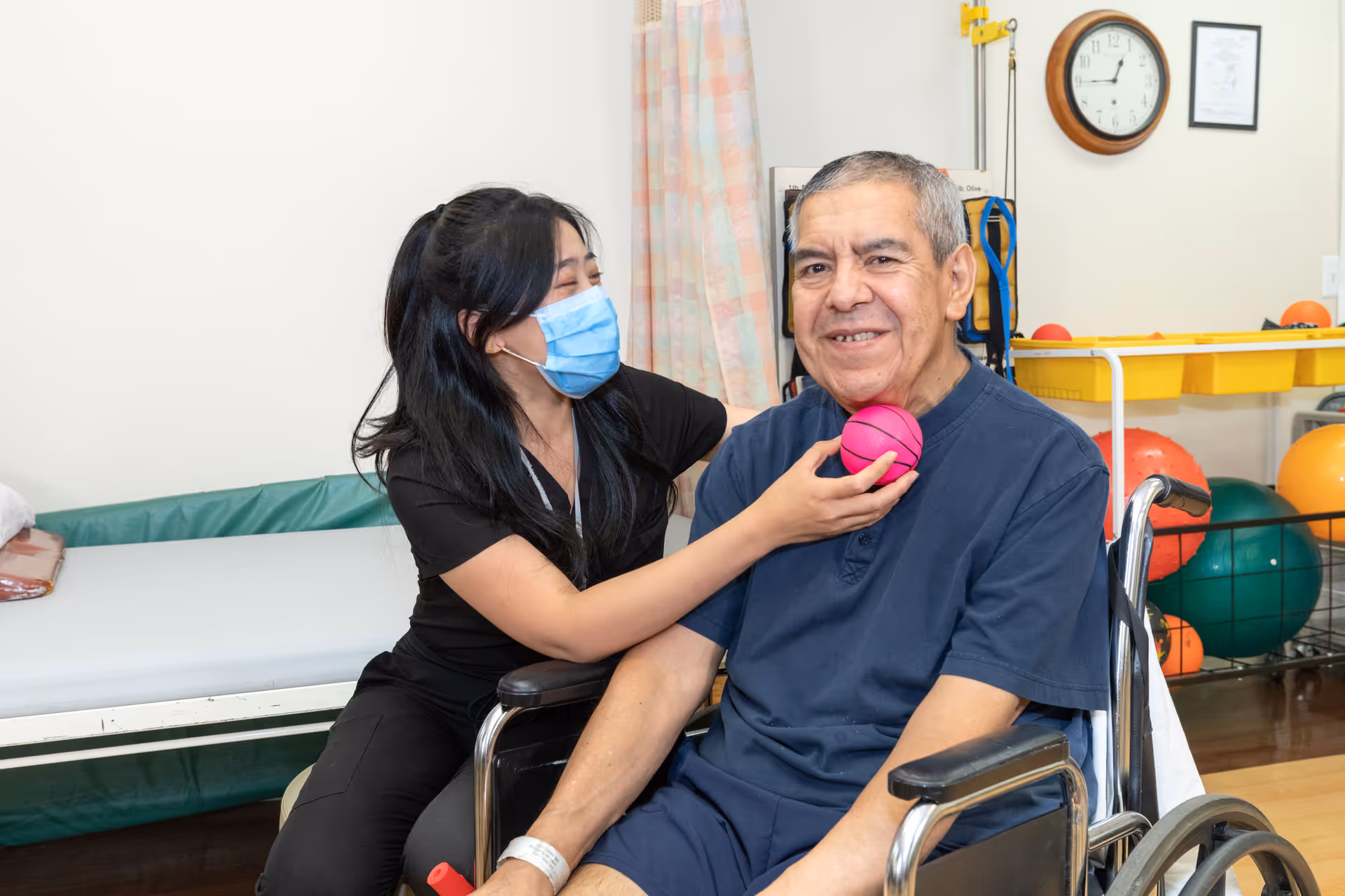 A healthcare worker wearing a face mask assists an elderly man in a wheelchair by holding a small pink exercise ball near his face in a therapy room with exercise equipment and a clock on the wall.