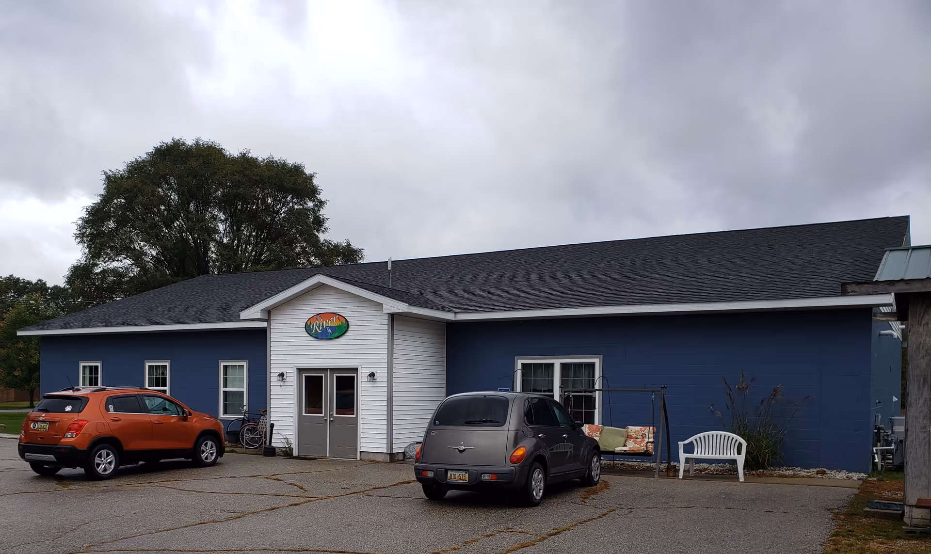 Exterior view of a single-story building with blue walls and a white entrance section. Two cars are parked in front of the building, and there is a swing bench and a white plastic chair near the window. A large tree is visible behind the building under a cloudy sky.