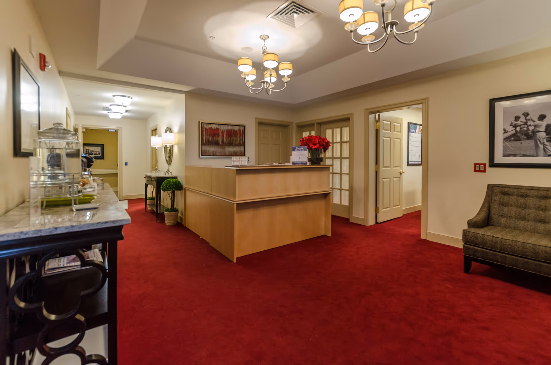Interior view of a senior living facility reception area with red carpet flooring, a wooden reception desk adorned with a vase of red flowers, a gray upholstered chair, framed artwork on the walls, and ceiling light fixtures.