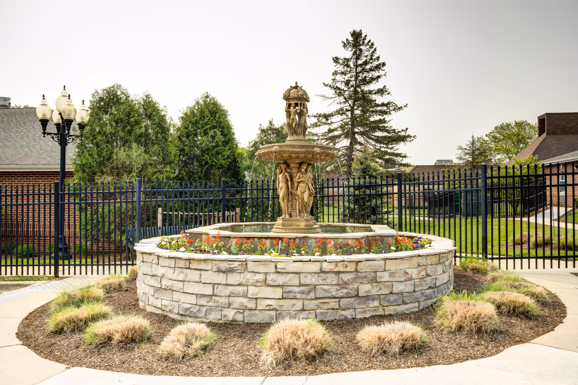 A decorative stone fountain with sculpted figures in the center, surrounded by a circular stone planter with colorful flowers. The fountain is located outdoors with a black metal fence, green trees, and brick buildings in the background under a clear sky.
