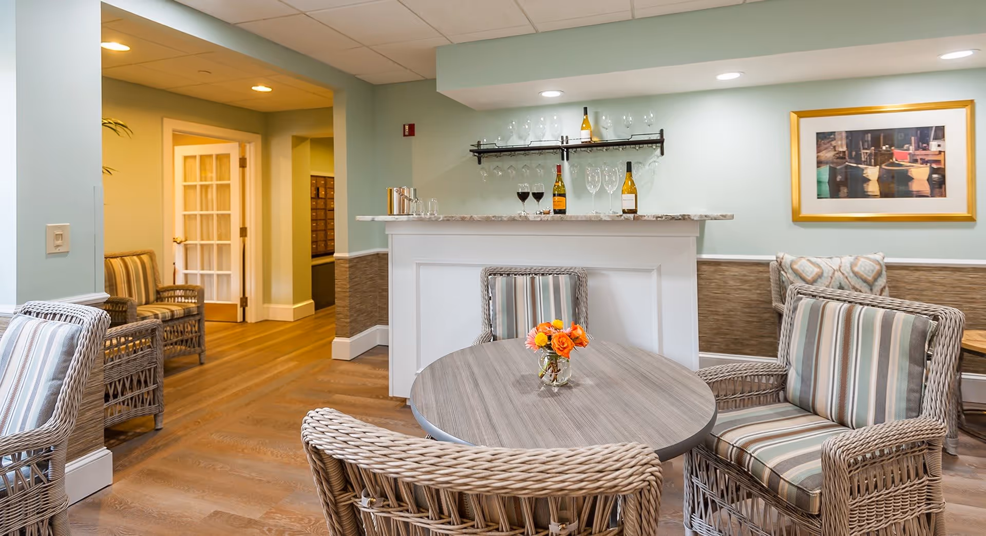 A cozy seating area in a senior living facility with a round wooden table surrounded by wicker chairs with striped cushions. A small vase with orange flowers is on the table. In the background, there is a white bar counter with wine bottles and glasses on a shelf above it. The walls are painted light blue with a framed picture hanging on the right side. The floor is wooden, and there is a hallway leading to other rooms.