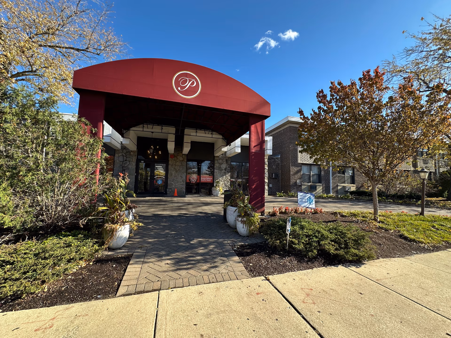 Entrance of Peterson Park Health Care Center with a maroon canopy featuring a circular logo with the letter 'P'. The building has stone and brick exterior walls, surrounded by landscaped bushes and trees with autumn foliage under a clear blue sky.