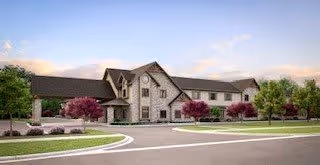 Exterior view of a two-story senior living facility building with a stone and siding facade, surrounded by landscaped greenery and trees under a partly cloudy sky.