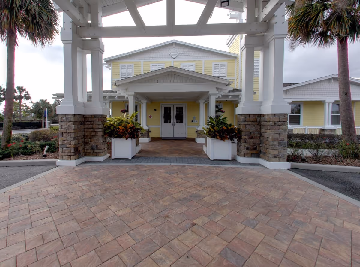 Front entrance of a yellow building with a covered portico supported by stone-clad columns, potted plants, and double doors.