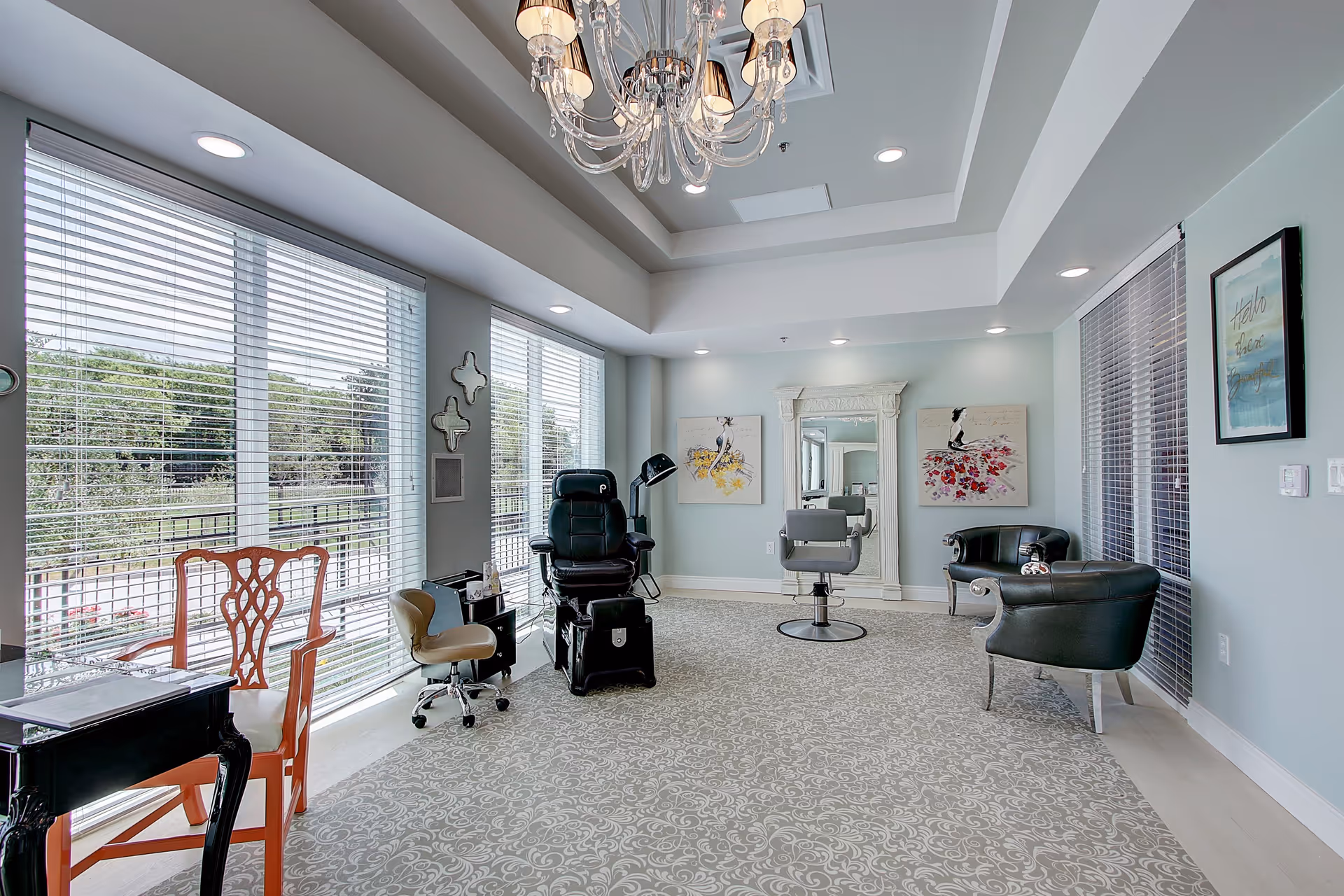 A bright and spacious salon area with large windows covered by blinds, featuring a black salon chair, a gray styling chair in front of a large ornate mirror, two black leather armchairs, a wooden chair, and a small black table. The room has light blue walls, a patterned carpet, and a chandelier hanging from the ceiling. There are two framed artworks on the wall and a framed quote on another wall.