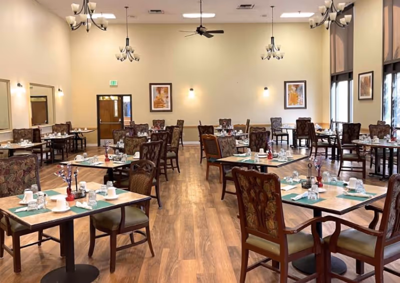 A spacious dining room with multiple square tables set with cups, glasses, napkins, and silverware. The room has wooden flooring, upholstered chairs, large windows with shades, chandeliers, ceiling fans, and framed artwork on the walls.