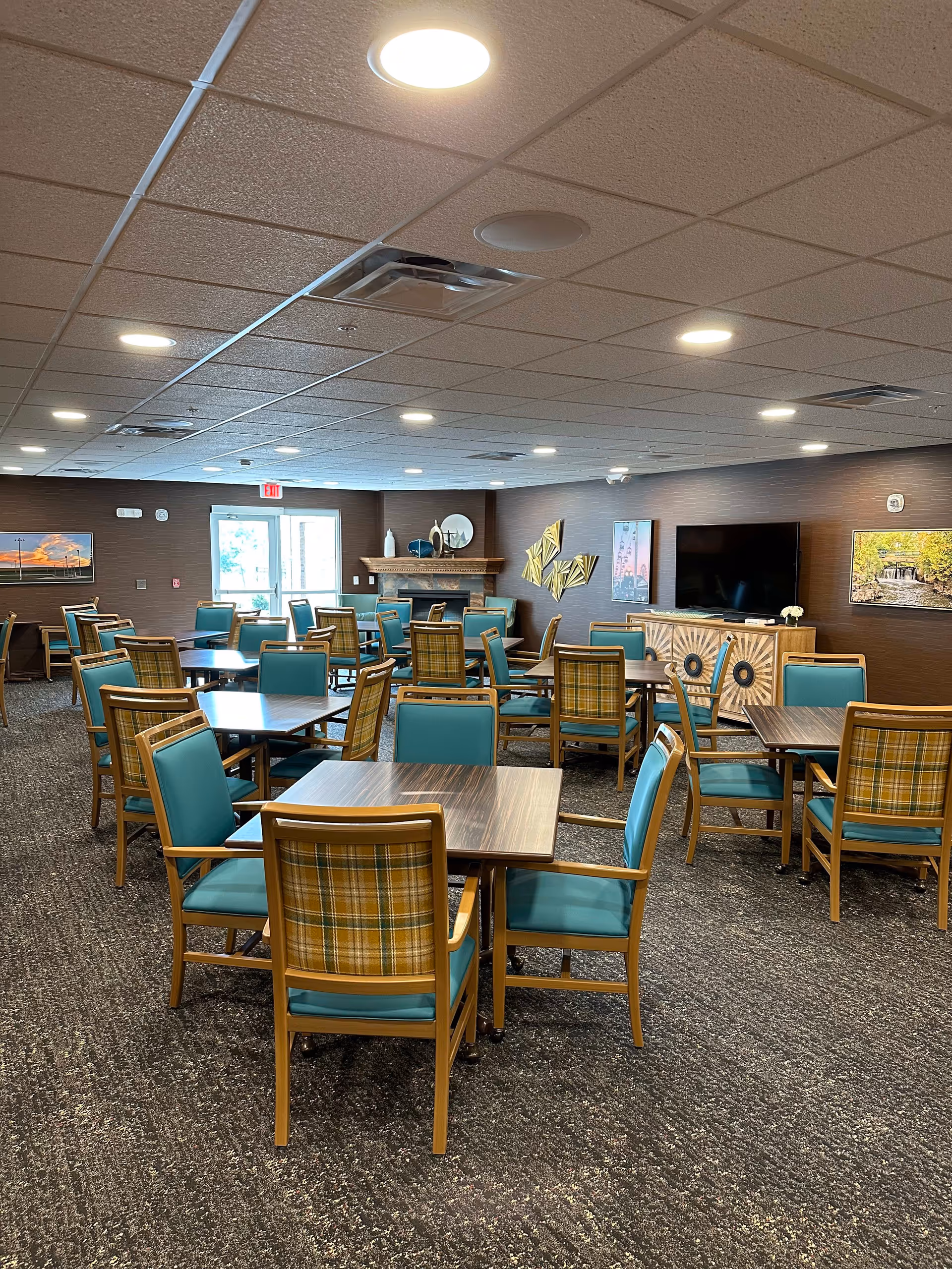 A spacious dining room with multiple wooden tables and chairs featuring teal cushions and plaid backs. The room has a carpeted floor, a ceiling with recessed lighting, and walls decorated with framed pictures and a TV mounted above a wooden cabinet. There is a fireplace in the back corner and a glass door letting in natural light.