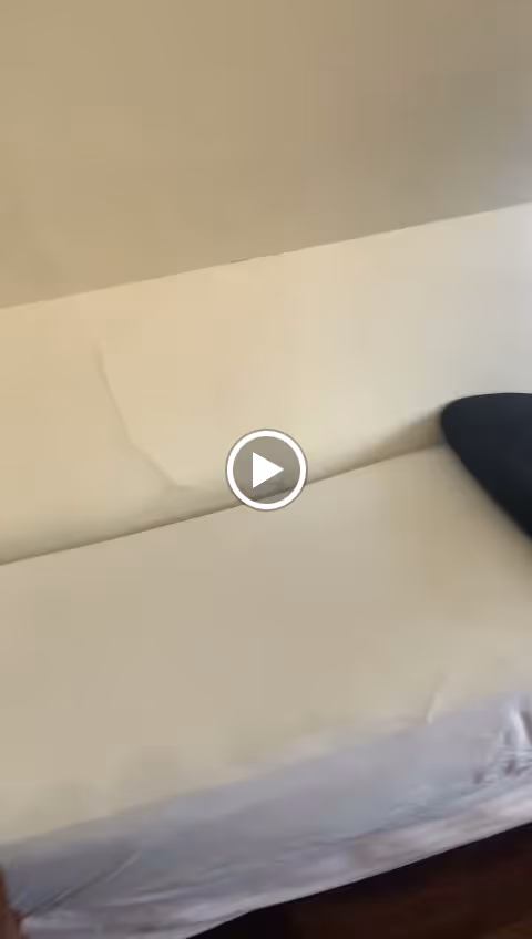 A simple white bed with a black pillow against a plain beige wall in a minimally decorated room.