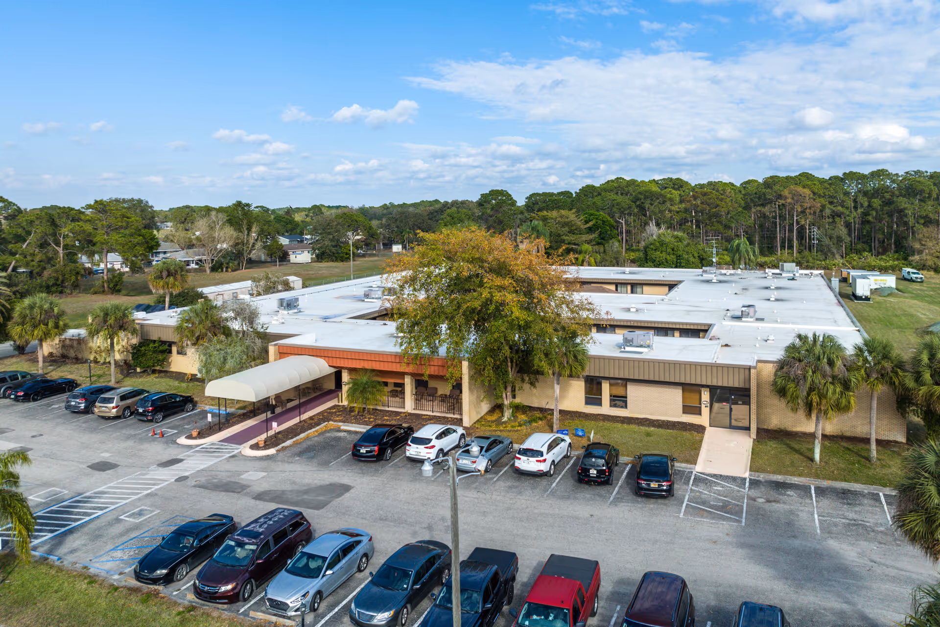 Aerial view of North Port Rehabilitation and Nursing Center showing a single-story building with a flat white roof surrounded by trees and greenery. There is a parking lot in front with several cars parked, and a covered entrance with a walkway leading to the building. The sky is partly cloudy with blue patches.