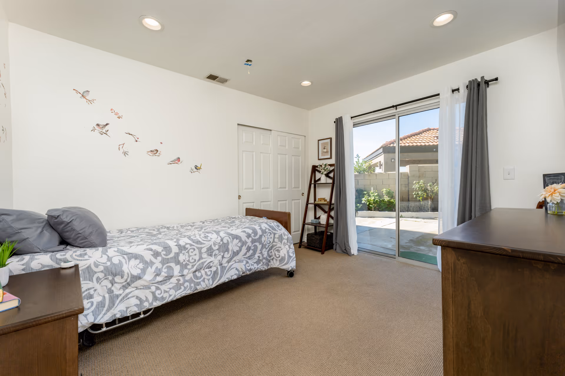 A bedroom with a single bed covered in a gray and white patterned bedspread, two gray pillows, and a small bedside table with a plant and books. The room has beige carpet, white walls with bird decals, a wooden dresser, a small ladder shelf with decor, and a sliding glass door leading to an outdoor patio area with a brick wall and plants.