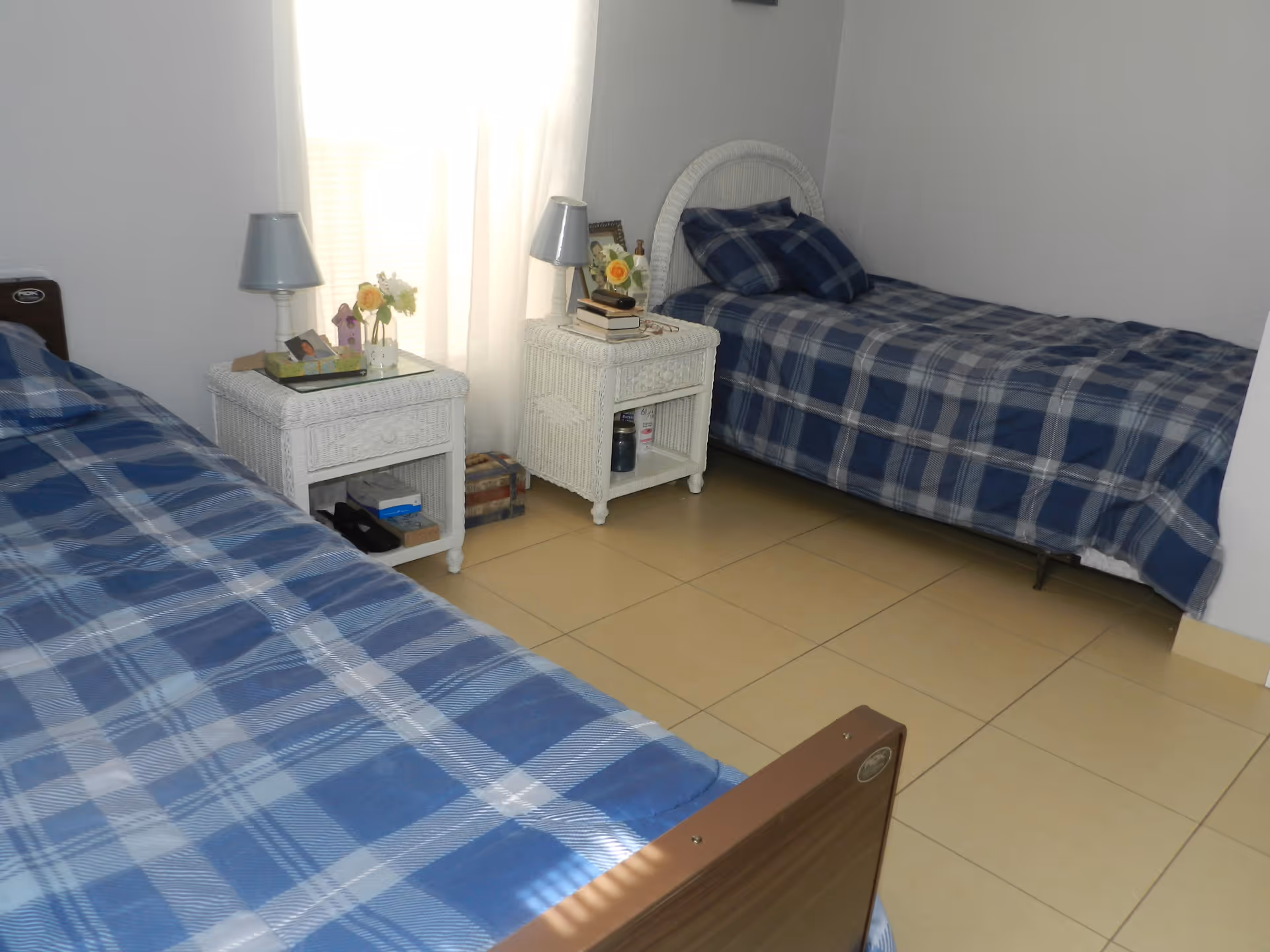 A bedroom with two single beds covered in blue plaid bedding. Between the beds are two white wicker nightstands, each with a small lamp and decorative items on top. A window with white curtains is in the background, allowing natural light into the room.