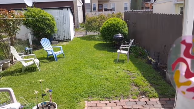 A small backyard garden area with green grass, several plastic chairs in white and blue, a black charcoal grill, and some potted plants. There is a wooden fence on the right side and a white shed at the back of the yard.