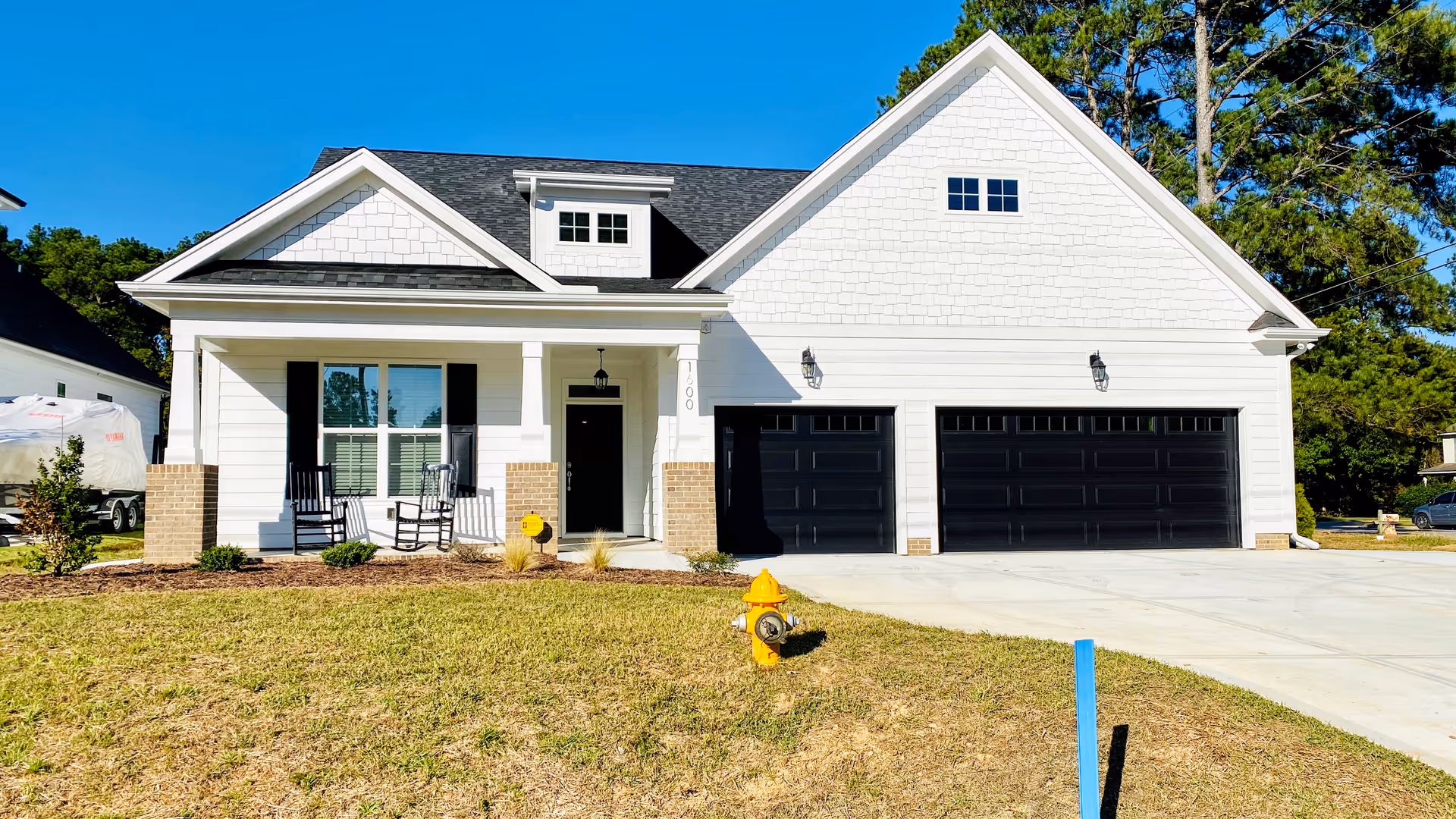 Front exterior view of a modern single-story white house with black shutters and a black front door. The house features a covered porch with two rocking chairs, a three-car garage with black doors, and a well-maintained lawn with a yellow fire hydrant near the driveway.
