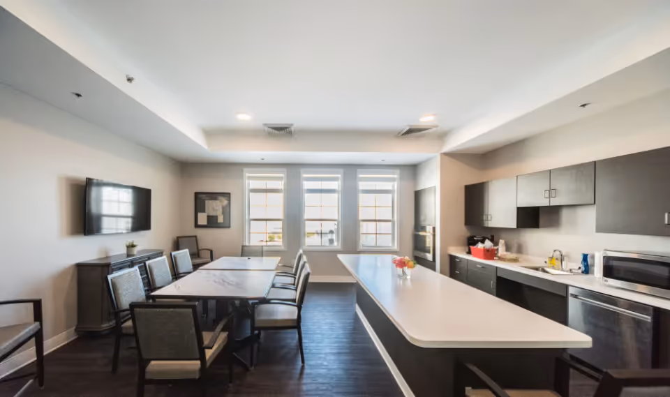 Bright communal dining area with a long table and chairs opposite a large island and kitchenette with cabinets and appliances.