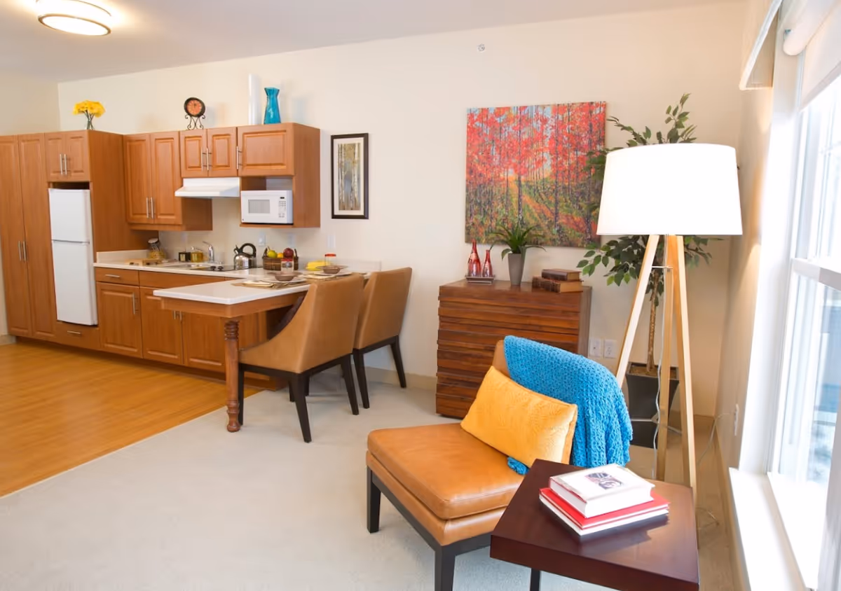Open-plan living area with a compact kitchen and breakfast bar, a leather lounge chair with colorful throw and pillow, floor lamp, and wall art.