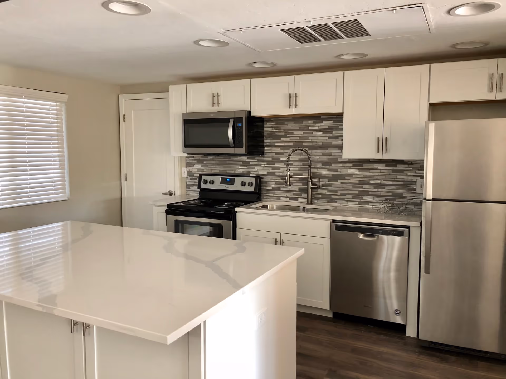 Modern kitchen with white cabinets, a stainless steel refrigerator, dishwasher, stove, and microwave. The backsplash features gray and white tiles, and there is a large white island countertop in the foreground. A window with blinds is on the left wall.