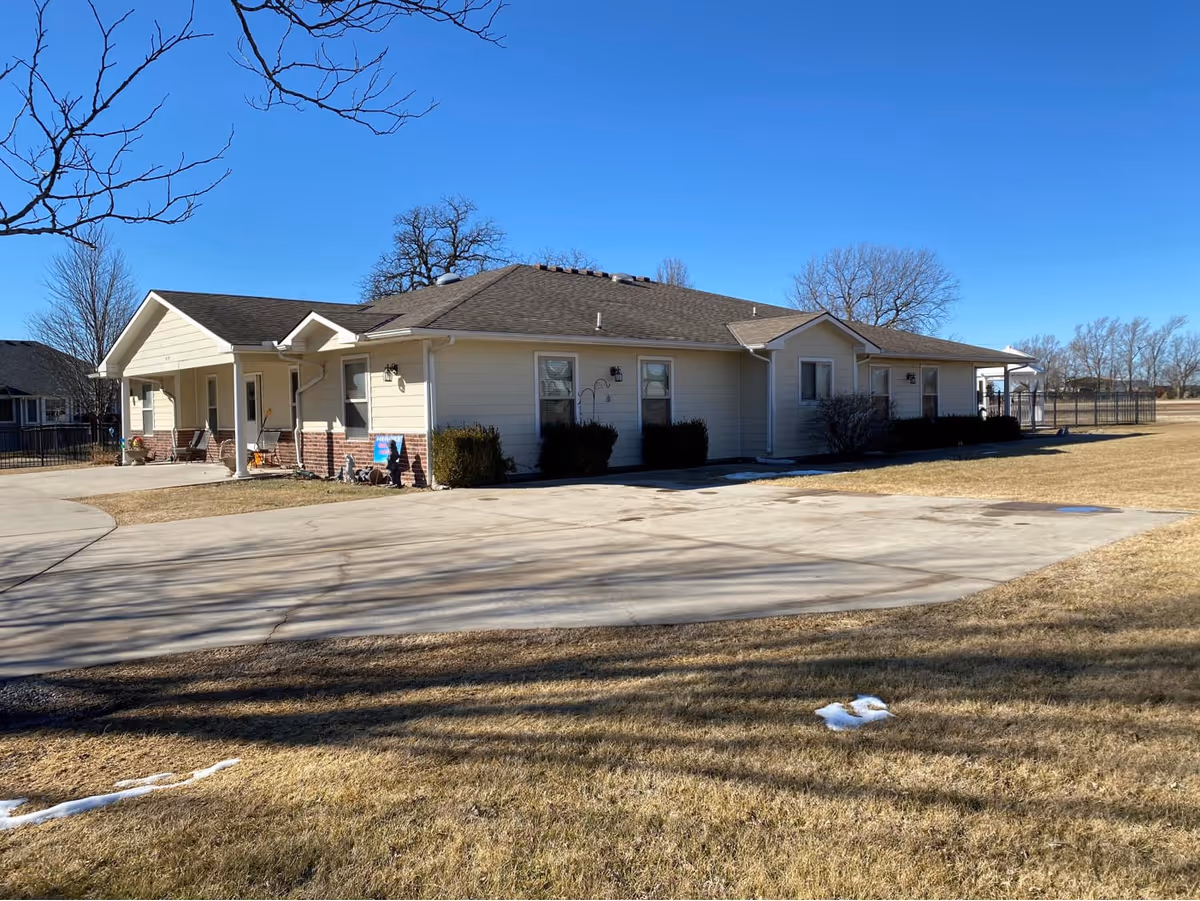 Single-story residential building with beige siding and a brown roof, surrounded by a large grassy area and a concrete driveway under a clear blue sky.
