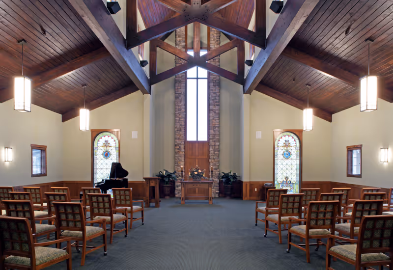 A chapel-like interior with a wooden vaulted ceiling, rows of chairs, stained-glass windows, a piano, and an altar at the far end.