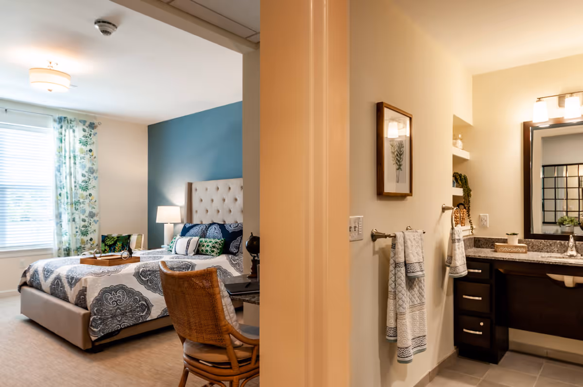 View of a bedroom and bathroom separated by a wall. The bedroom features a bed with a tufted headboard, patterned bedding, a bedside lamp, a window with floral curtains, and a wicker chair at a small desk. The bathroom has a granite countertop with a sink, a large mirror, wall-mounted light fixtures, and towels hanging on a rack.