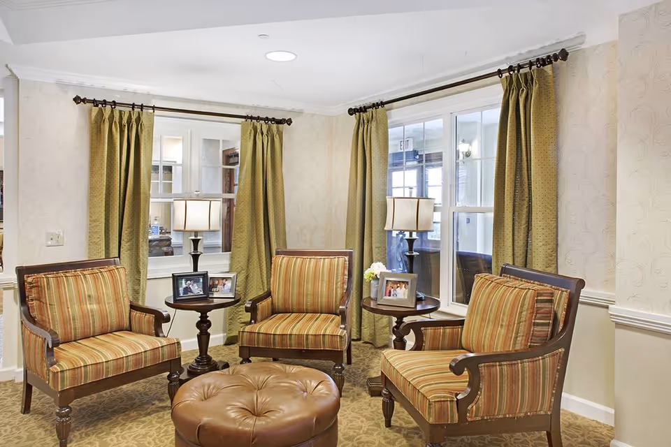 Cozy sitting area with three striped armchairs, round side tables with lamps and framed photos, and a tufted leather ottoman in front of windows with green curtains.
