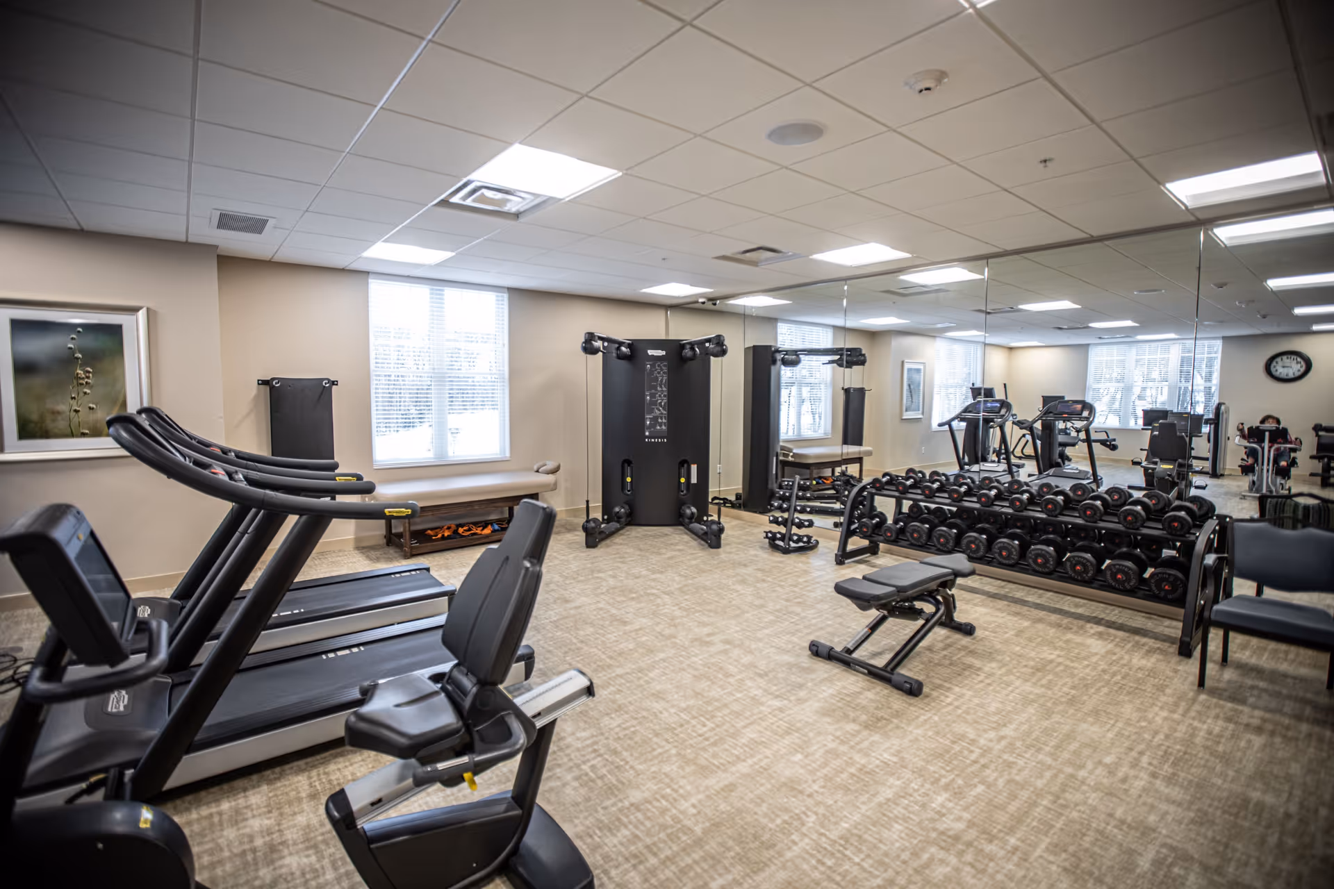 A bright fitness room with treadmills, a recumbent bike, free weights, exercise bench, and mirrored wall.