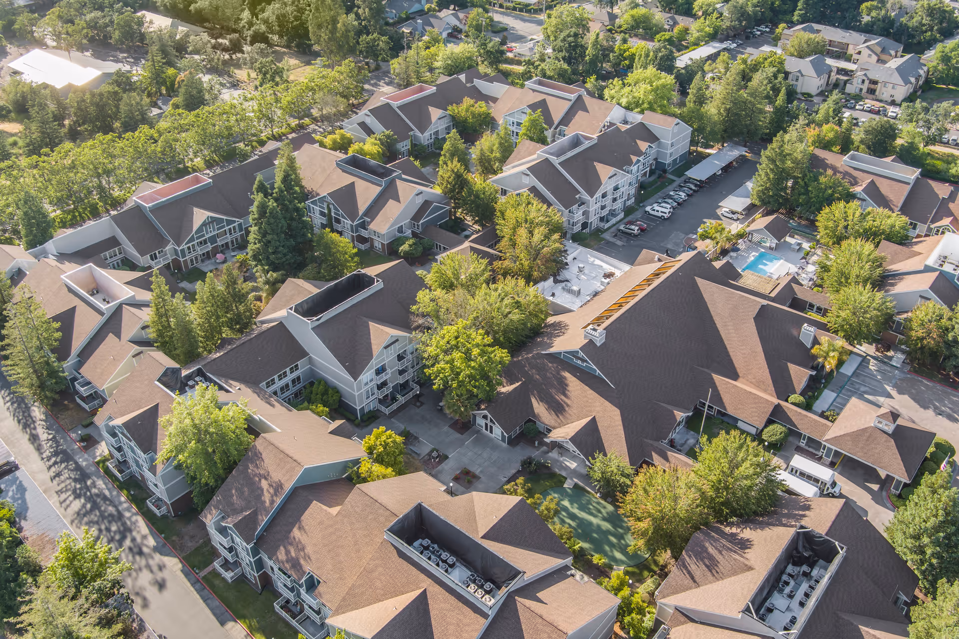Aerial view of a senior living facility with multiple buildings surrounded by trees and greenery. The buildings have brown roofs and light-colored walls, with parking areas and a swimming pool visible within the complex.