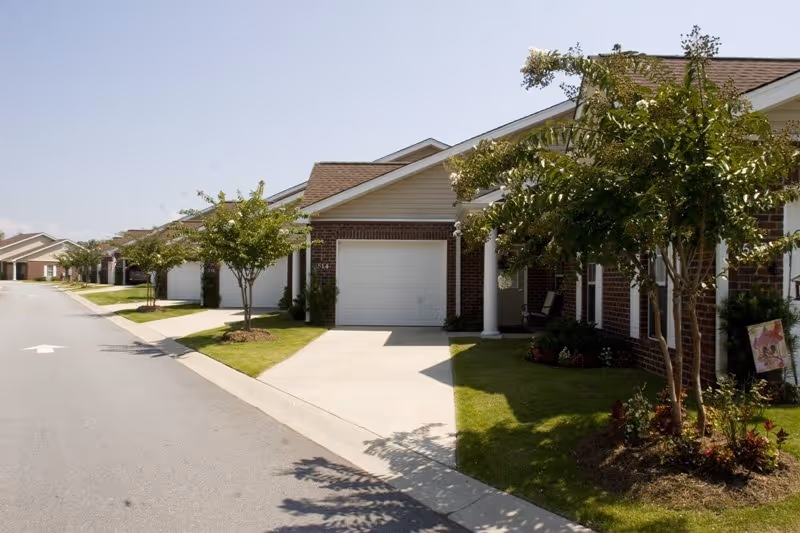 View of a row of single-story residential buildings with garages, small trees, and well-maintained lawns along a paved street under a clear sky.