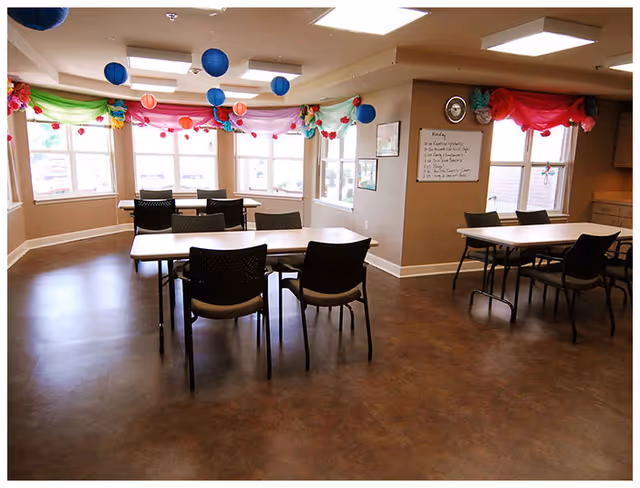 Well-lit communal dining/activity room with tables and chairs, windows, and colorful hanging decorations.