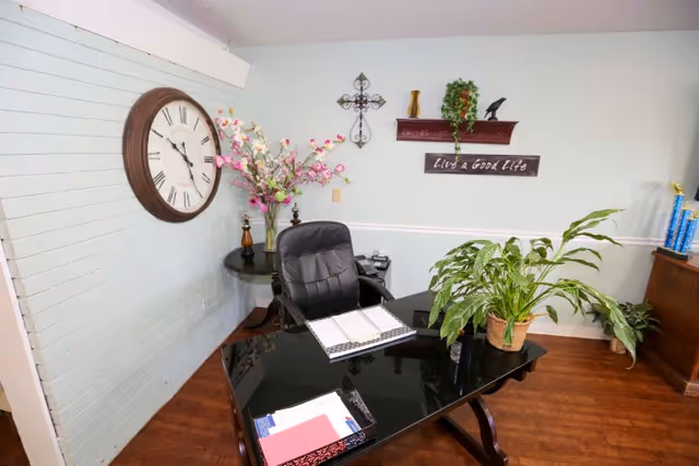 An office area with a black glass desk, a black office chair, a large wall clock, a vase with pink flowers on a round side table, a potted plant on the desk, and wall decor including a cross and a sign that reads 'Live a Good Life'.