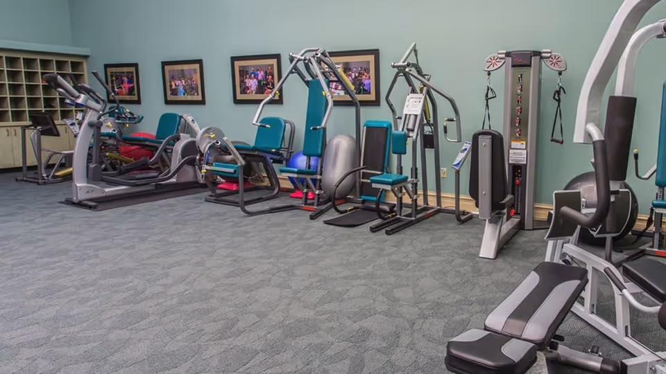 A fitness room with various exercise equipment including treadmills, weight machines, and resistance training machines. The room has a light blue wall with framed pictures and a carpeted floor.