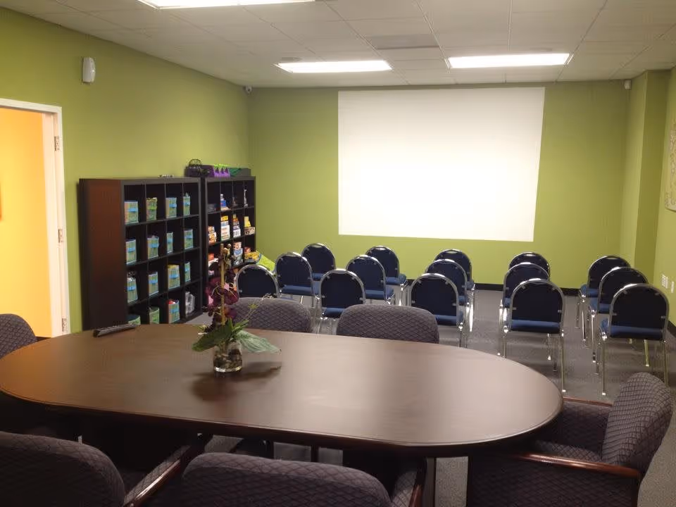A room with a large oval wooden table surrounded by cushioned chairs in the foreground and rows of blue chairs facing a blank white projection screen on a green wall in the background. There is a black shelving unit with various items on the left side and a small plant centerpiece on the table.