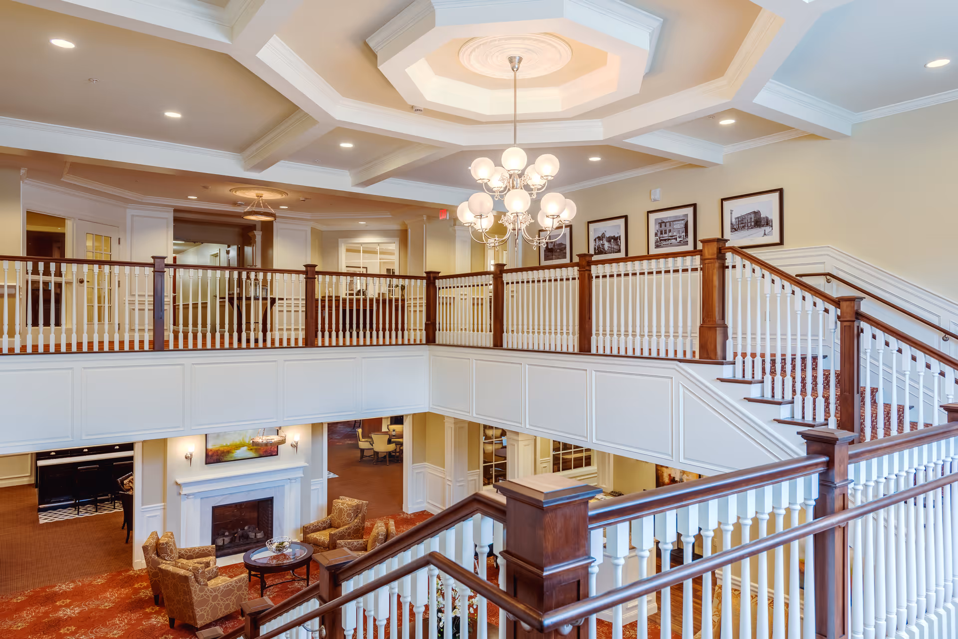 Interior view of a senior living facility featuring a two-story open area with a wooden staircase and white railings. The lower level has a seating area with armchairs around a fireplace, and the upper level has framed pictures on the wall. A chandelier hangs from the ceiling, illuminating the space.
