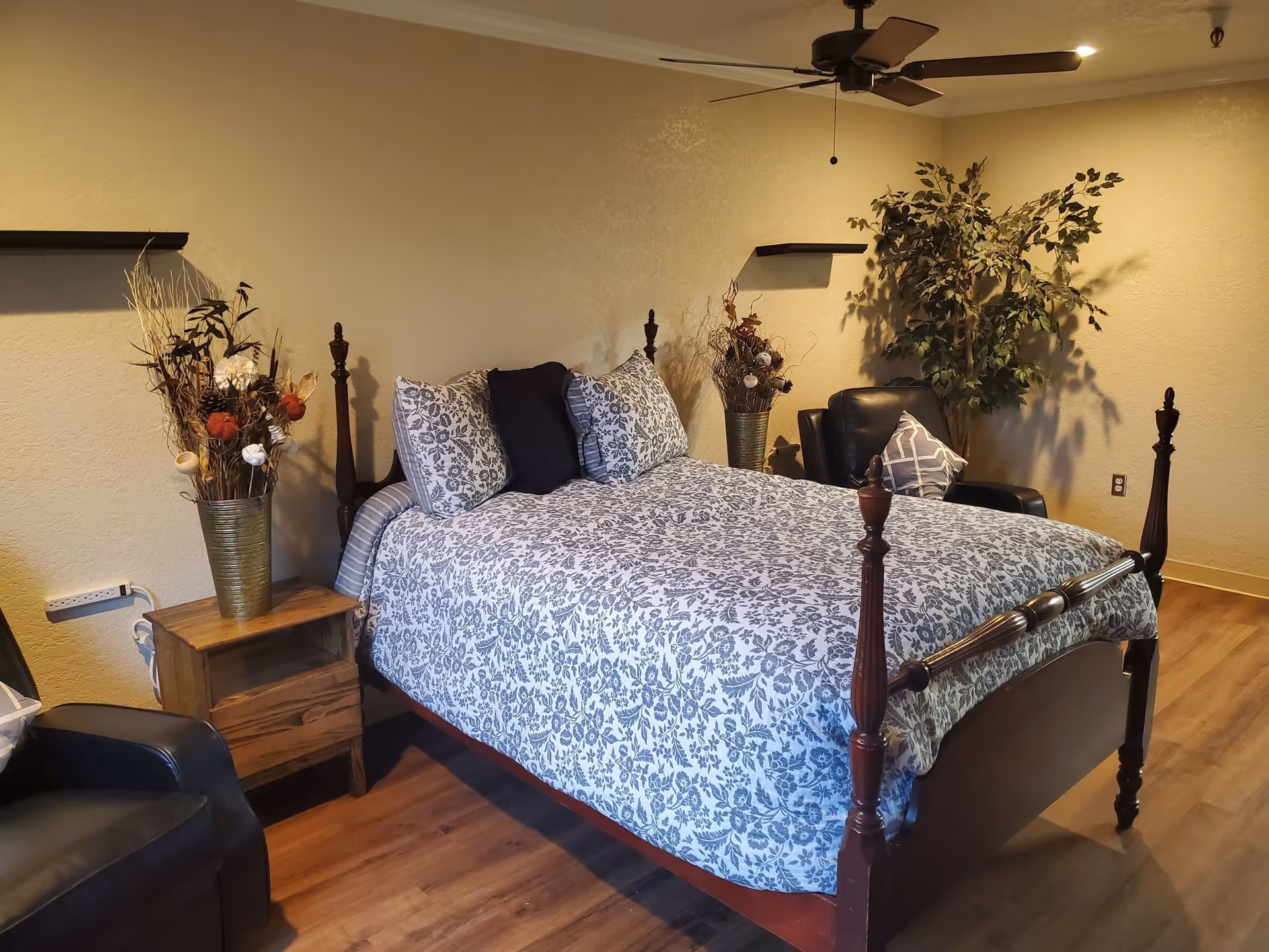 Cozy bedroom with a four-poster bed dressed in patterned bedding, flanked by a wooden nightstand, armchairs, a potted plant and ceiling fan.