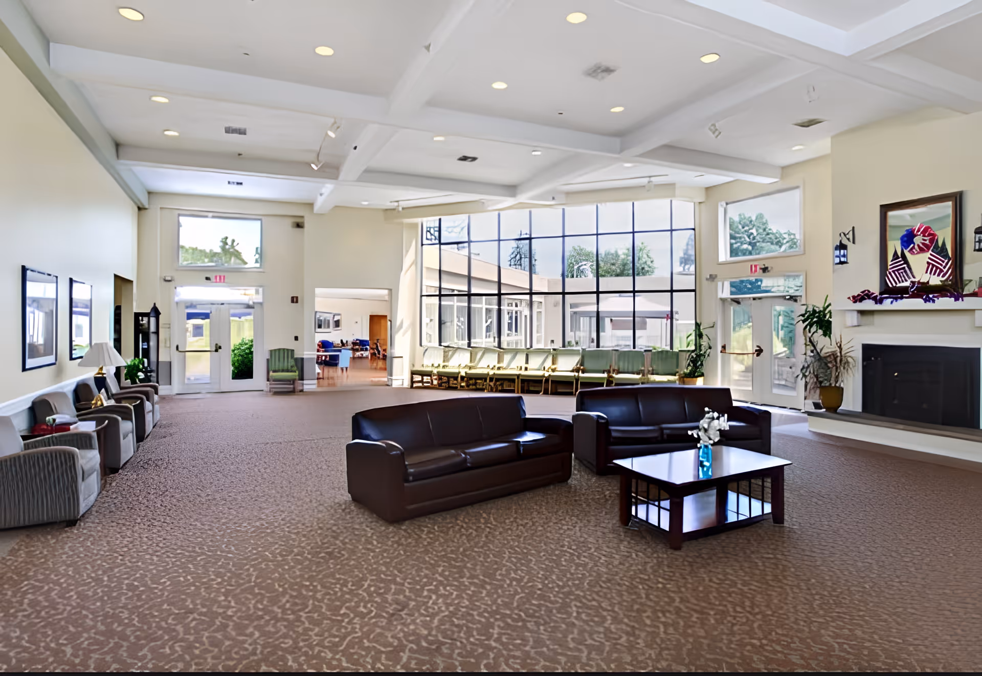 Spacious senior living facility common area with large windows allowing natural light, two dark brown leather sofas around a wooden coffee table with a small flower vase, multiple chairs lined up against the walls, a fireplace decorated with a framed picture and ornaments, and doors leading to other rooms.