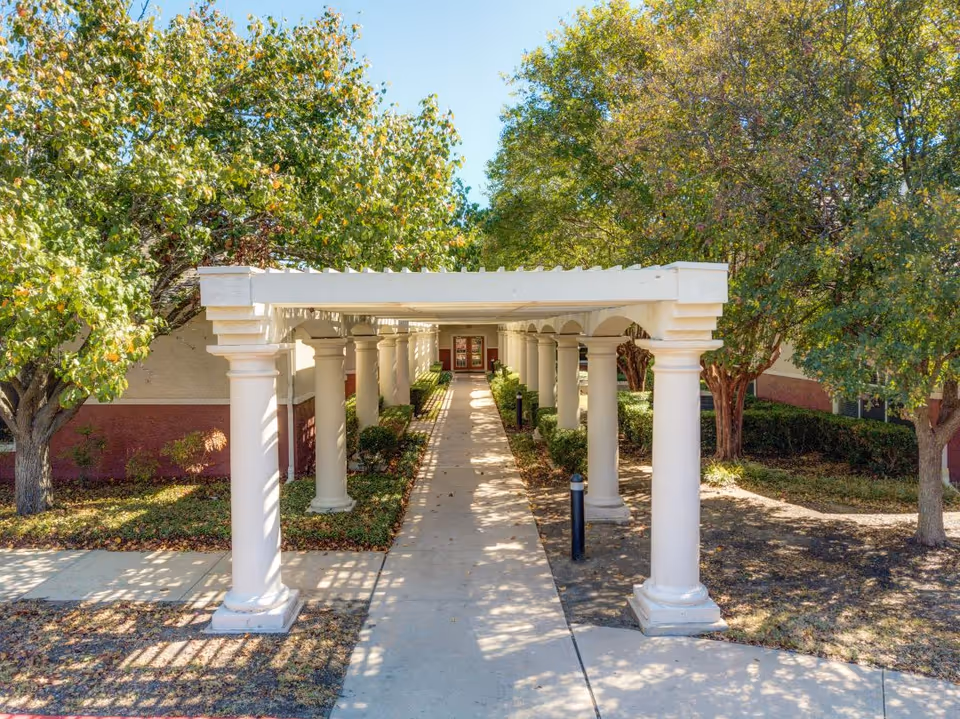 A covered walkway with white columns on both sides leading to a building entrance, surrounded by trees with green leaves and some fallen leaves on the ground.