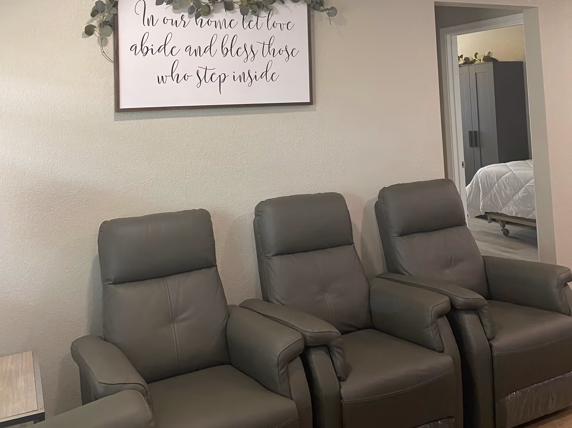 Three gray recliner chairs sit against a wall beneath a framed script sign, with a doorway showing a bedroom.