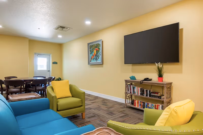 A bright common area with a blue sofa and green armchairs facing a wall-mounted TV above a small bookshelf, with a dining table visible in the background.